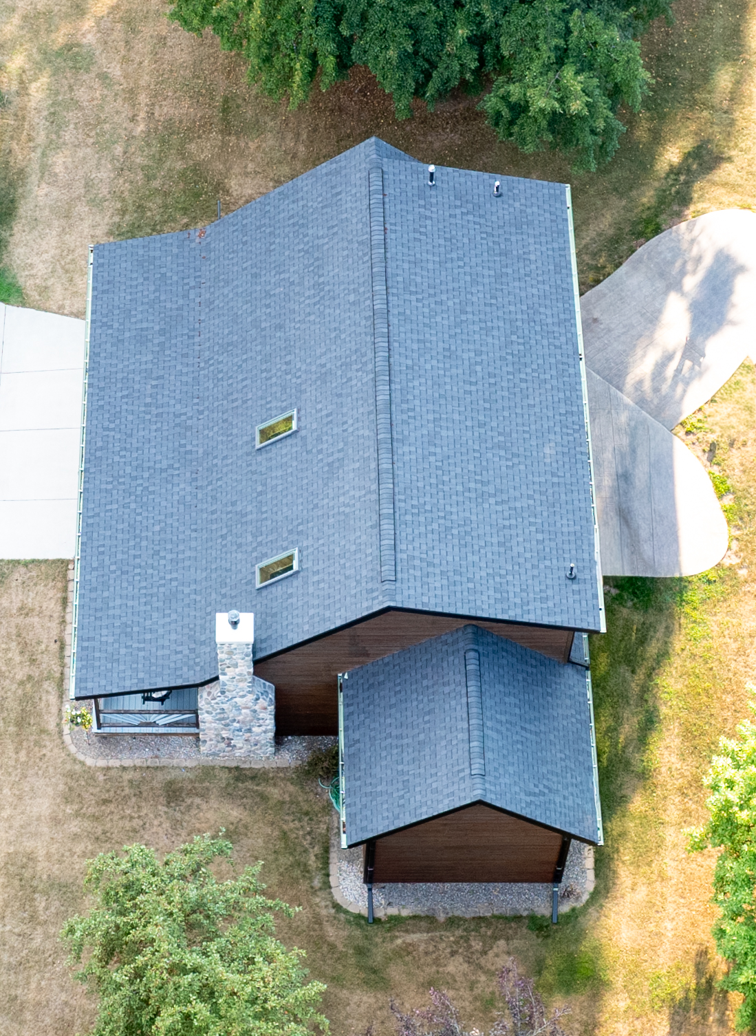Aerial view of a steep architectural shingle roof with skylights, stone chimney and covered entry